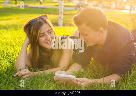 Portrait ensoleillée de sweet young couple lying détente sur l'herbe et écoute de la musique des écouteurs sur smartphone, journée d'été. Banque D'Images