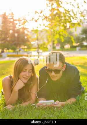 Portrait ensoleillée de sweet young couple lying détente sur l'herbe et écoute de la musique des écouteurs sur smartphone, journée d'été. Banque D'Images