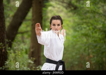 Jeune femme pratiquant des mouvements de karaté dans son bois de forêt - Kimono Blanc - Ceinture Noire Banque D'Images