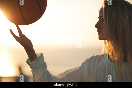 Jeune femme debout devant un coucher du soleil la rotation d'un terrain de basket-ball sur son doigt. Banque D'Images