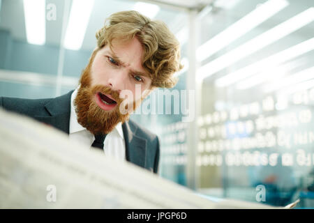 Portrait de barbu choqué businessman reading newspaper in office, fronçant avec bouche ouverte Banque D'Images