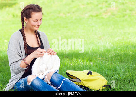 Une jeune femme aux cheveux noirs tressés en amorces en haillons tricots jeans avec des aiguilles à tricoter de la laine pull vert thread, assis sur un brillant gre Banque D'Images