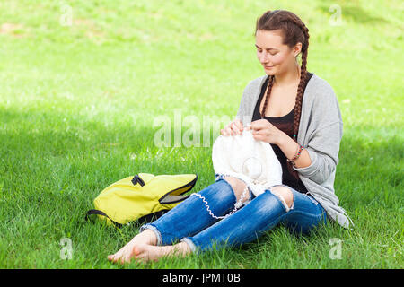 Jeune femme aux cheveux noirs tressés en amorces dans des jeans déchirés tricote avec des aiguilles à tricoter de la laine pull vert thread assis sur un vert lumineux gr Banque D'Images