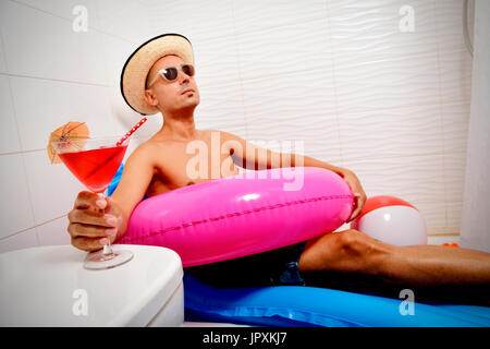 Un jeune homme de race blanche portant des lunettes de soleil, un chapeau de paille et un anneau de bain rose prétendre qu'il est relaxant dans un matelas d'air bleu placé sous la douche d'un Banque D'Images