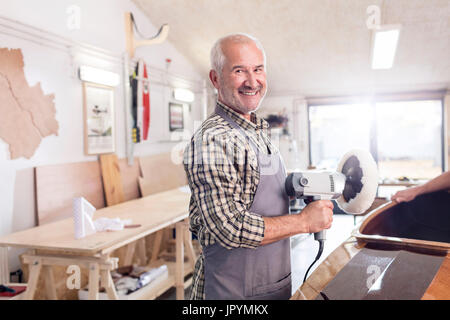 Portrait souriant, confiant senior male carpenter en utilisant un tampon sander sur bateau en bois dans l'atelier Banque D'Images