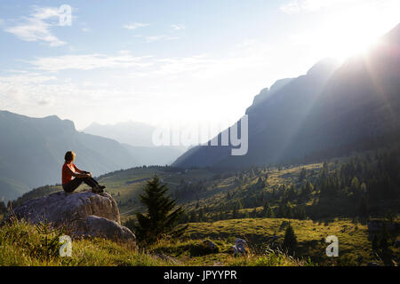 Femme en tee-shirt orange assis sur un rocher et profiter en vue imprenable au coucher du soleil, des montagnes de Montasio, Alpes Juliennes, Frioul-Vénétie Julienne, Italie. Banque D'Images