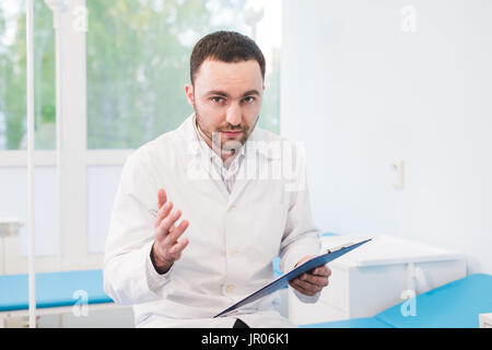 Cheerful young doctor holding un presse-papiers et des gestes avec sa main à l'hôpital Banque D'Images