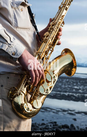 Un homme se tient avec un saxophone sur la plage au bord de l'eau ; l'Alaska, États-Unis d'Amérique Banque D'Images