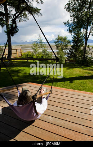 Woman Relaxing In Hammock sur une terrasse en bois avec vue sur l'océan et les montagnes ; Alaska, États-Unis d'Amérique Banque D'Images