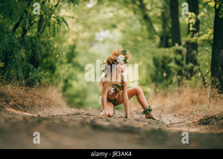 Belle petite fille en image de nymphe dryade et avec tête floral wreath siège en route forestière. Banque D'Images