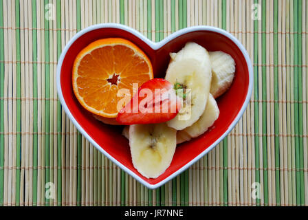 Vibrant, contraste élevé de fruits (mandarine, fraise, banane) dans un plat en forme de coeur vert placé sur un tapis de bambou et neutre. Banque D'Images