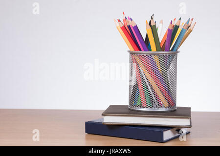 Collection de crayons de couleur dans un panier métallique en haut d'une pile de livres scolaires ou de revues sur un bureau bureau en bois avec copie espace sur le mur Banque D'Images