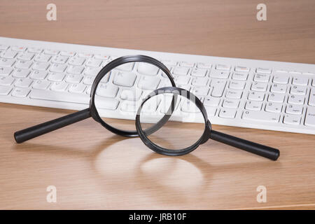 Deux loupes en équilibre sur le bord d'une table en bois ou un bureau avec un clavier d'ordinateur dans un concept de recherche et d'analyse en ligne Banque D'Images