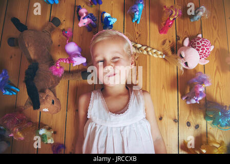 Sourire et clignant de petite fille couchée dans ses jouets. L'intérieur horizontal tourné. Banque D'Images