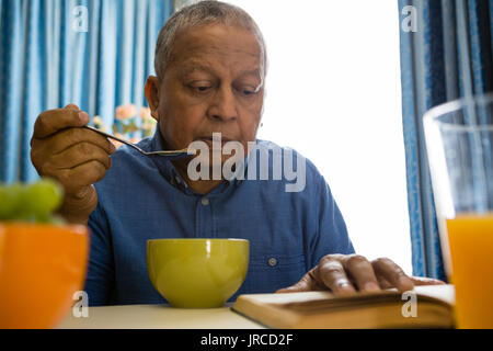 Senior ayant l'alimentation lors de la lecture de livre à table dans la maison de soins infirmiers Banque D'Images