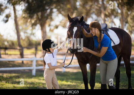 Happy girl et femme jockey cheval caresser en se tenant sur le terrain au paddock Banque D'Images