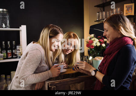 Une jeune femme montrant quelque chose sur smartphone pour deux soeurs jumelles blondes dans le café. Trois femmes à la recherche d'amis quelque chose sur le téléphone, boire du café un Banque D'Images