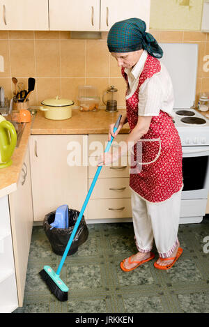 Une femme adulte, une femme au foyer ou d'une femme de chambre, portant un tablier et un foulard vert sur sa tête qui balayent la cuisine avec un balai Banque D'Images