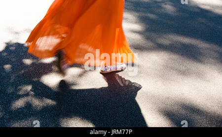 Les jambes d'une jeune femme marche dans la longue robe orange palpitations dans summer breeze avec trottoir tree shadows Banque D'Images