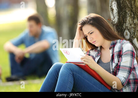 Seul triste student looking at n'a pas de séance d'examen sur l'herbe dans un parc avec des gens à l'arrière-plan flou Banque D'Images