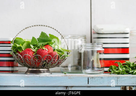 Fraisiers jardin frais mûrs et Melissa herbes dans vase vintage debout avec verre vide et metal pots pour confiture sur la table de cuisine en bois blanc bleu. Banque D'Images