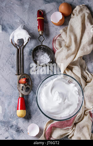 Ingrédients et ustensiles pour la cuisson de la meringue. Les blancs d'oeufs fouettés, le sucre en poudre, oeufs cassés, vintage et tamis mélangeur de main sur lin gris serviette sur te Banque D'Images