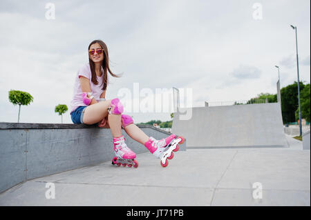 Portrait d'une jolie jeune femme en short, t-shirt, lunettes de soleil et de rollers assis sur le banc en béton dans la piscine patinoire roller Banque D'Images