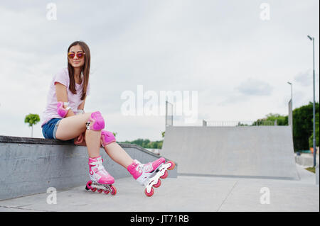 Portrait d'une jolie jeune femme en short, t-shirt, lunettes de soleil et de rollers assis sur le banc en béton dans la piscine patinoire roller Banque D'Images