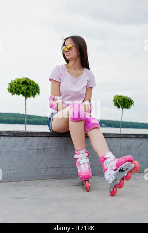 Portrait d'une jolie jeune femme en short, t-shirt, lunettes de soleil et de rollers assis sur le banc en béton dans la piscine patinoire roller Banque D'Images