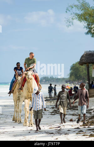 Deux touristes à cheval sur des chameaux sur la plage avec des gens marcher le long de leur côté, Diani, Kenya Banque D'Images