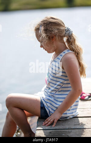 Petite fille assise sur une plate-forme de bois par la rivière ou du lac tandis que les projections d'eau par les pieds. Banque D'Images