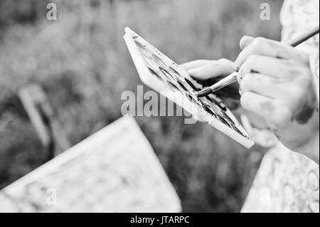 Photo en gros plan de mains tenant des peintures à l'aquarelle et un pinceau tandis que la peinture dans la nature. Photo en noir et blanc. Banque D'Images