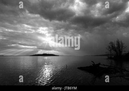 Les rayons du soleil émergeant dans les nuages au-dessus d'une île sur un lac, avec des arbres et des faisceaux dans l'avant-plan Banque D'Images