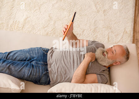 Surpris homme barbu senior using digital tablet while sitting on sofa at home Banque D'Images