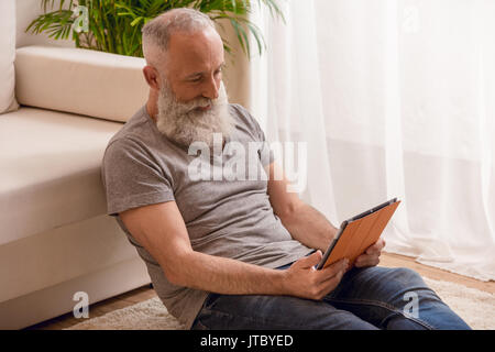 Homme barbu senior smiling and using digital tablet et assis sur le plancher à la maison Banque D'Images