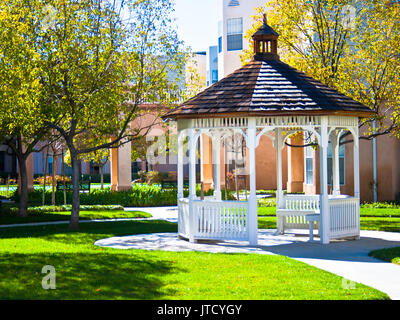 Un gazebo blanc brille dans une cour ensoleillée entourée d'herbe et les arbres feuillus. Banque D'Images