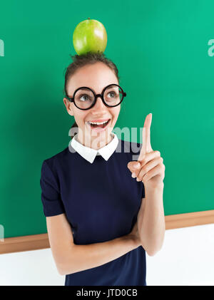 Joyeux élève enthousiaste avec apple sur sa tête un doigt pointé vers le haut. Photo de teen à lunettes près de tableau noir, concept créatif avec retour à la sc Banque D'Images
