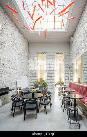 Restaurant avec des murs texturés et une grande fenêtre au plafond. Il y a des sofas rouges et gris, de tables rondes en bois, tabourets et chaises, plantes, Banque D'Images