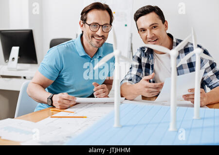 Les travailleurs enthousiastes. Sourire agréable hommes assis à la table et à la station d'énergie éolienne du modèle sur la table Banque D'Images