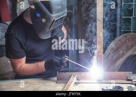 Close-up un jeune homme chauve dans un masque noir et t-shirt noir une machine à souder à l'arc dans la construction d'un atelier sombre, autour d'un lot o Banque D'Images