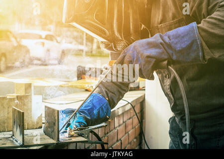 Soudeur en homme masque de soudage, de la construction et de gants de protection uniforme bleu brasse à souder sur métal de construction de la rue, dans l'arrière-plan un vieux b Banque D'Images