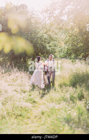 La jeune mariée dans la longue robe blanche et le marié sont habillés moderne portant le bicecle avec bouquet rose et de marche dans la forêt verte ensoleillée. Banque D'Images