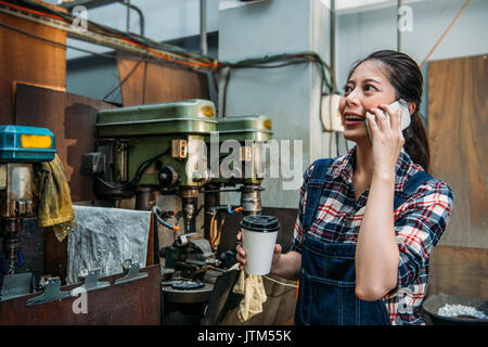 Le personnel féminin de la fraiseuse attrayant holding Coffee cup papier en position de repos et à l'aide de forage ministère téléphone intelligent d'appeler chatting Banque D'Images