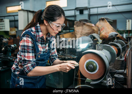 Tour attrayant travailleur féminin maintenir les composants métal et le port de lunettes de sécurité travaillant sur meule avec des étincelles dans machine-w Banque D'Images