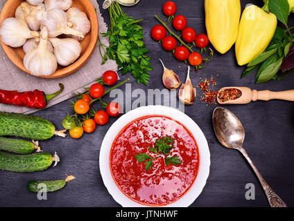 Tomate fraîche et soupe de légumes gaspacho dans une plaque ronde blanche avec une cuillère de fer sur un fond noir, autour les ingrédients et épices Banque D'Images