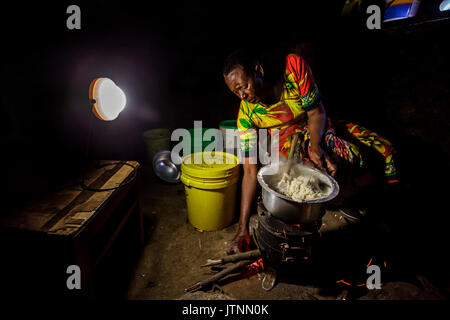 Chez elle, près d'Arusha, en Tanzanie, Julieth Mollel, sœur de Solar, prépare un dîner d'ugali, de légumes et de haricots sur sa cuisinière propre. L'ugali est un aliment de base consommé dans de nombreux pays d'Afrique et est de la bouillie de farine de maïs. Travailler dans sa cuisine extérieure compacte la nuit est plus facile maintenant à la fois avec la cuisinière propre qui émet très peu de fumée et utilise seulement une fraction du bois de chauffage d'une cuisinière traditionnelle à trois pierres, mais aussi avec sa lanterne solaire Solar Sister pour éclairer la zone de façon éclatante elle travaille. Banque D'Images