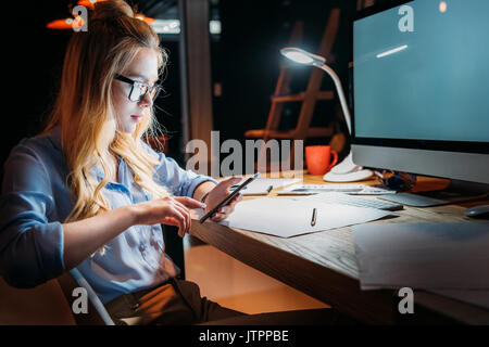 Jeune blond caucasian businesswoman à lunettes en utilisant smartphone et assis à lieu de travail avec ordinateur Banque D'Images