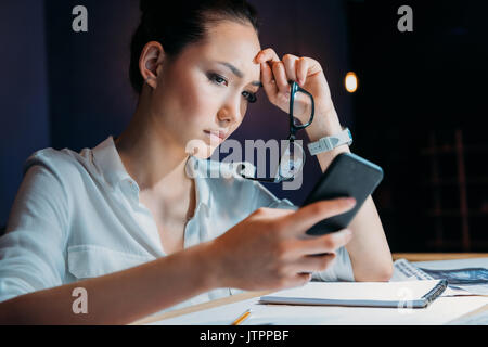 Pensive fatigué asian businesswoman holding eyeglasses and using smartphone, travailler tard au bureau Banque D'Images