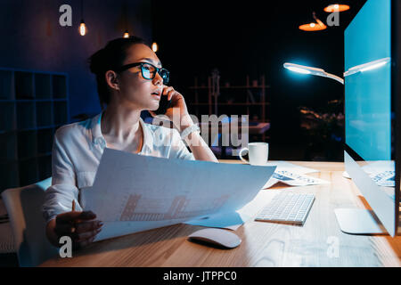 Young businesswoman in eyeglasses holding blueprint et parler sur smartphone Banque D'Images