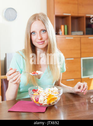 Happy blonde long-haired woman eating fruit salade avec du yogourt en home Banque D'Images